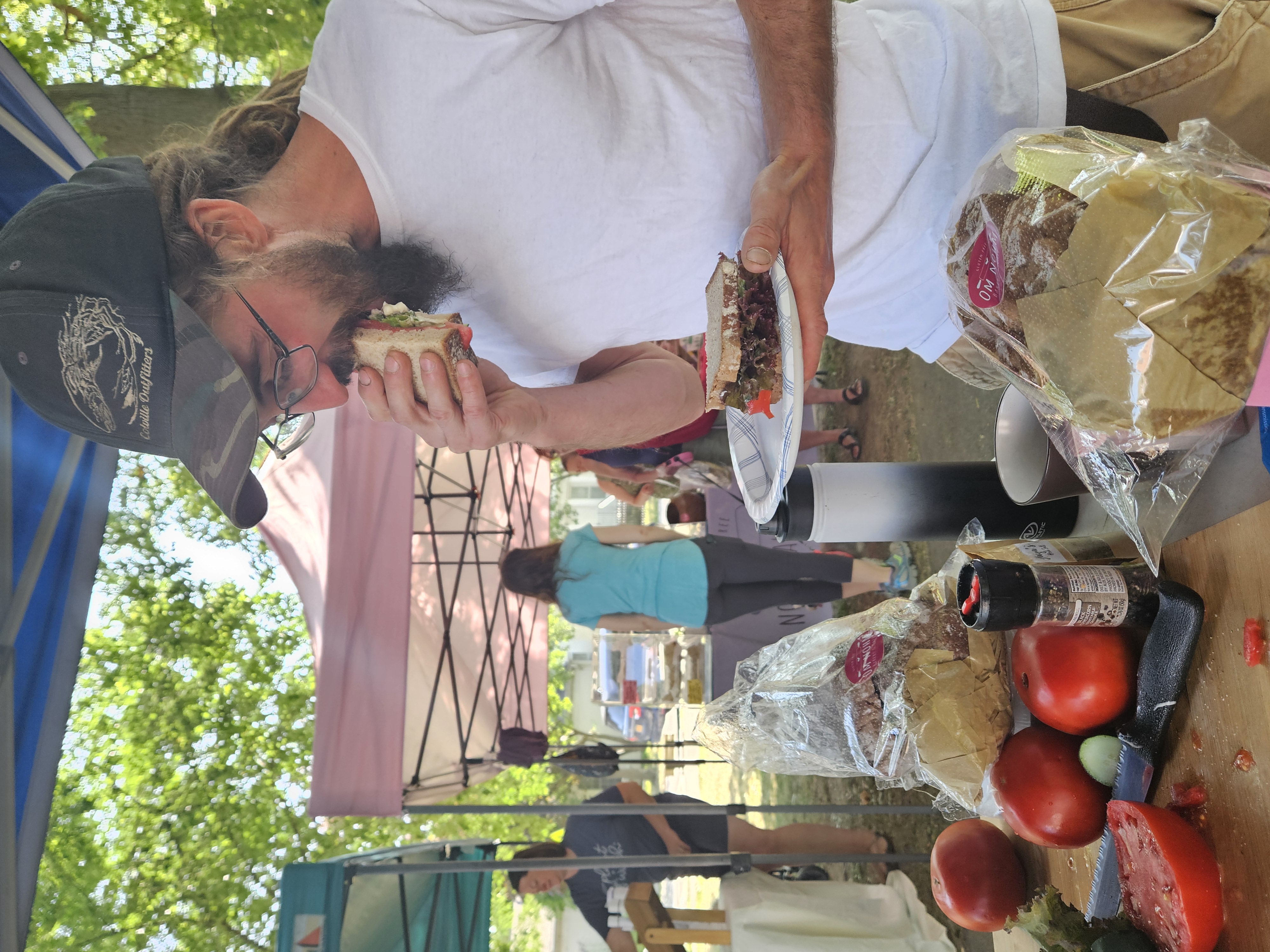 A market guest taking a hearty bite of a fresh sandwich under the vendor tents.