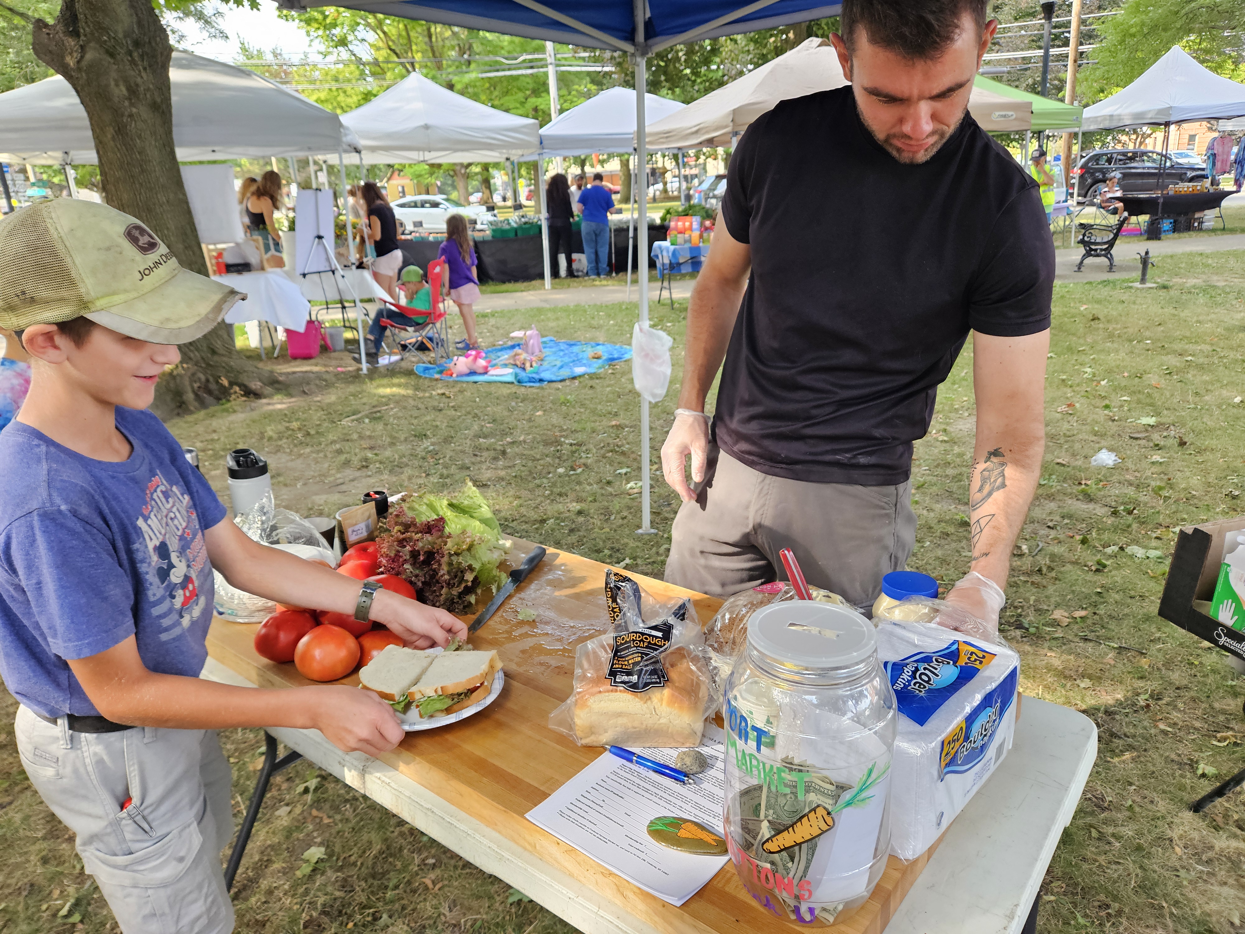 Donation jar on the table next to tomatoes, lettuce, and bread, with a young helper smiling.