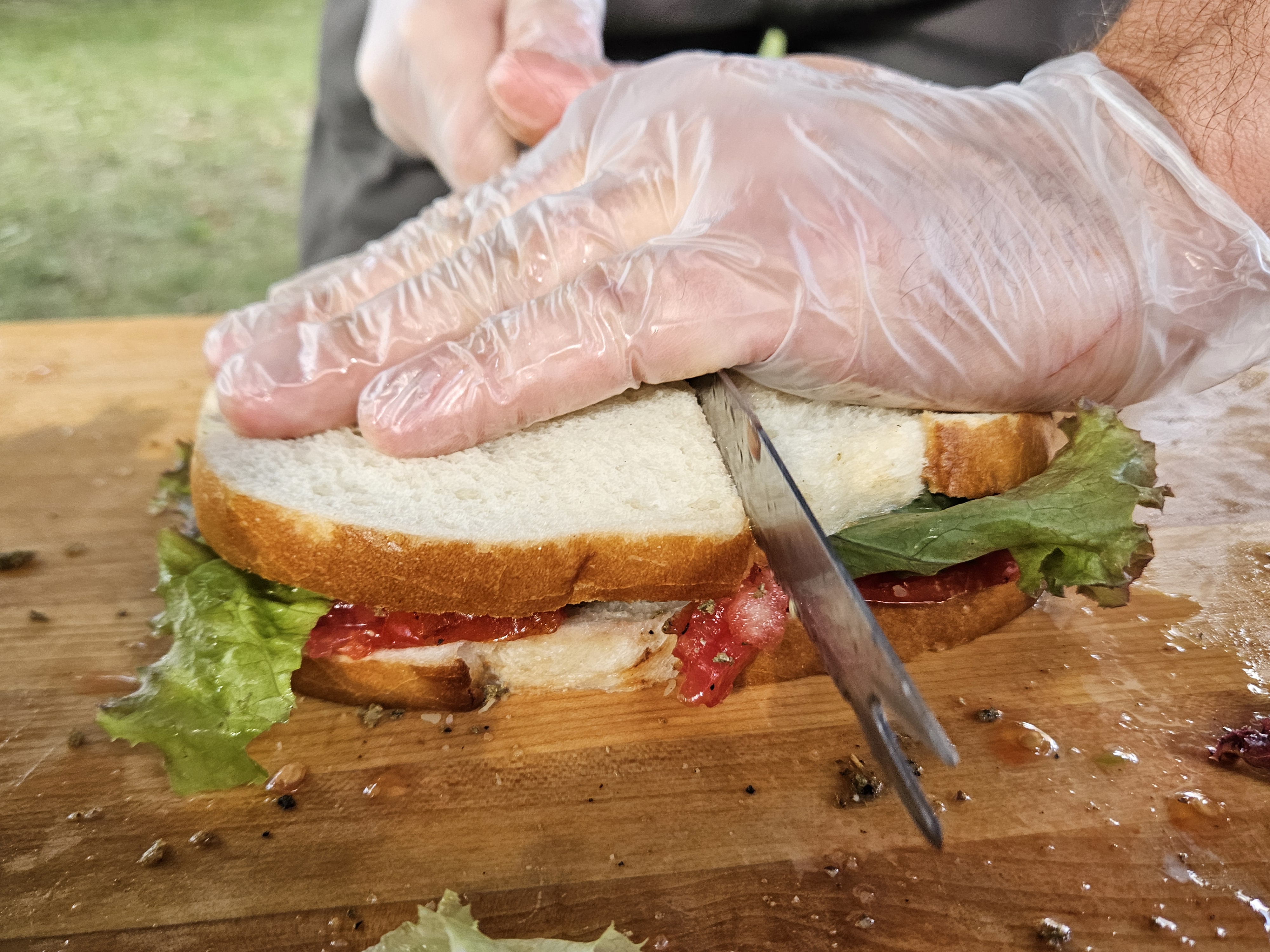 Gloved hands slicing a fresh tomato-lettuce sandwich on a maple butcher block.